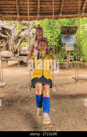 Karen Frau aus Pai in Mae Hong Son, Thailand, mit Messing Halsringen sitzt auf einer Holzschaukel im rustikalen Dorf und trägt ein farbenprächtiges Karen Kleid Stockfoto