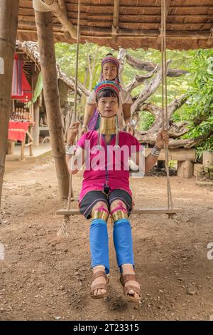 Karen Frau aus Pai in Mae Hong Son, Thailand, mit Messing Halsringen sitzt auf einer Holzschaukel im rustikalen Dorf und trägt ein farbenprächtiges Karen Kleid Stockfoto