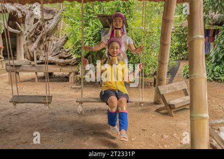 Karen Frau aus Pai in Mae Hong Son, Thailand, mit Messing Halsringen sitzt auf einer Holzschaukel im rustikalen Dorf und trägt ein farbenprächtiges Karen Kleid Stockfoto