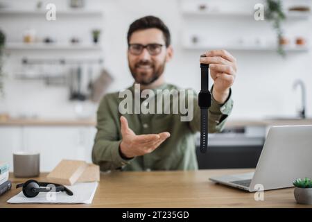 Verschwommener Hintergrund eines lächelnden bärtigen Bloggers, der im Home Office sitzt und eine neue gekaufte Smart Watch zeigt. Fokus auf neues modernes Gerät in der männlichen Hand. U Stockfoto