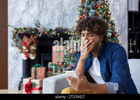 Langweiliger Mann, der zu Silvesterferien und Weihnachten auf dem Sofa sitzt, gähnender hispanischer Mann, der ein Smartphone im Wohnzimmer in der Nähe des Weihnachtsbaums hält. Stockfoto