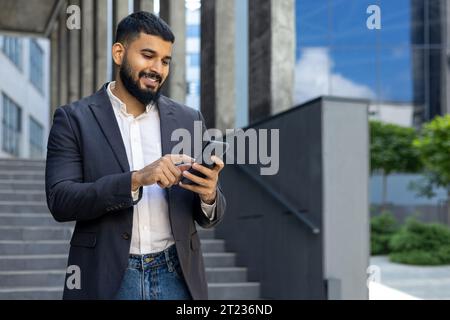 Lächelnder junger muslimischer Geschäftsmann, der das Telefon benutzt, während er vor einem Bürozentrum steht. Stockfoto
