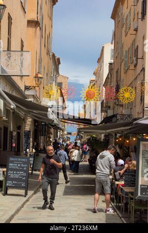 Frankreich, Korsika, l'Ile-Rousse, Rue Dominique Fioravanti: Einkaufslustige füllen die fröhlich geschmückte Gasse im Schatten der Markisen. Stockfoto