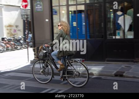 Eine Frau mit langen Haaren und Sonnenbrille in eleganten Casuals fährt durch eine Straße im Zentrum von Paris, Frankreich. Stockfoto