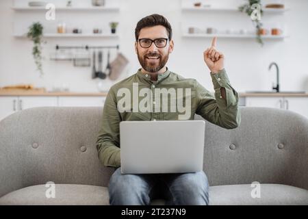 Glücklicher bärtiger Mann, der den Finger nach oben zeigt, während er auf einer bequemen Couch sitzt und einen tragbaren Laptop benutzt. Aufgeregtes männliches Gefühl, inspiriert durch neue Ideen Stockfoto