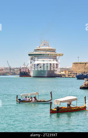Valletta, Malta - 3. August 2023: Kleine Holzboote fahren Touristen über den Hafen in der Stadt Valletta. Im Hintergrund befindet sich ein Kreuzfahrtschiff Stockfoto