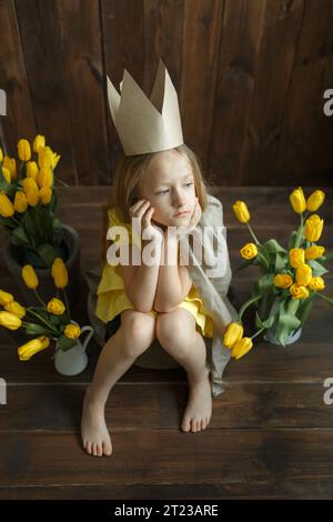 Porträt einer traurigen Prinzessin, die neben gelben Tulpensträußen sitzt. Sie sitzt traurig und denkt nach. Vertikaler Rahmen. Stockfoto