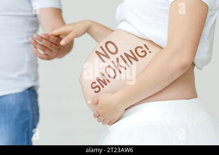 Eine schwangere Frau hält die Hand ihres Mannes am Bauch mit der Inschrift - RAUCHEN VERBOTEN, Ausrufezeichen. Schwangerschaftskonzept. Stockfoto