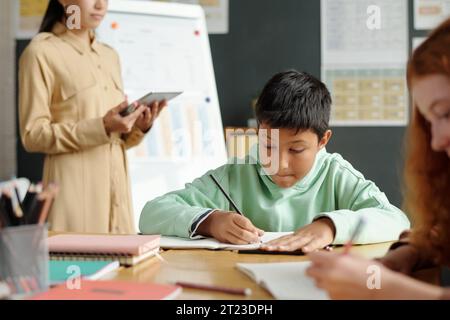 Konzentriere dich auf niedliche multiethnische Schuljungen in Casualwear, die englische Grammatikregeln im Copybook aufschreiben, während sie unter Lehrer und Klassenkameraden sitzen Stockfoto