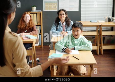 Glücklicher multiethnischer Schuljunge und zwei Schulmädchen, die den Lehrer anschauen und neue Grammatikregeln erklären, während sie vor dem Publikum stehen Stockfoto