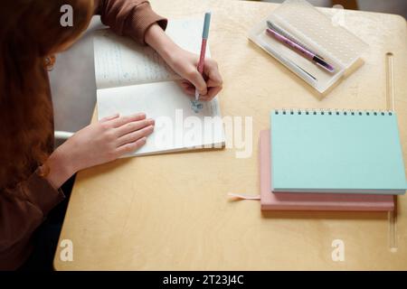 Über dem Winkel der Hände des Schülers der Sekundarstufe, der in einem Copybook mit Stift zeichnet, während er am Schreibtisch sitzt, während er am Unterricht des Englischgrammatikkurses sitzt Stockfoto