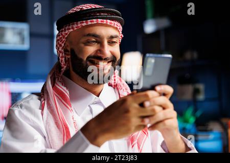 Ein lächelnder arabischer Mann sitzt an einem Schreibtisch und surft mit seinem Smartphone im Internet. Selektiver Fokus auf einen enthusiastischen islamischen Kerl in traditioneller Kleidung, der sein digitales Mobilgerät greift. Stockfoto