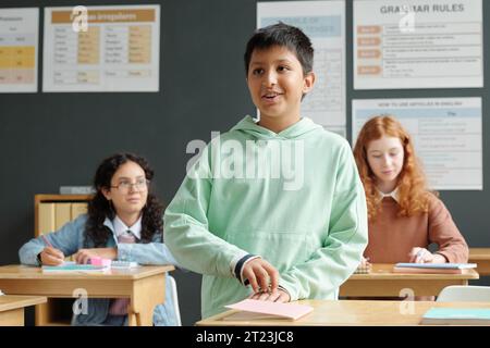 Glücklicher Schuljunge im Kapuzenpullover, der am Schreibtisch steht und die Frage des Lehrers beim Unterricht der englischen Grammatik gegen zwei Schulmädchen beantwortet Stockfoto