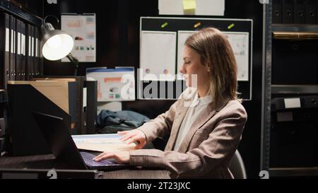 Eine Privatdetektivin steht und verlässt das Büro mit ihrem Rucksack. Die kaukasische Polizistin schließt ihren drahtlosen Computer und bereitet ihren Schreibtisch vor, bevor sie für den Tag aufbricht. Stockfoto