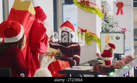 Überfröhlicher afroamerikanischer Arbeiter, der Geschenke vom Weihnachtsmann in Weihnachtsarbeitsräumen erhält. Ein aufregender Mitarbeiter bekommt Geschenke von einem verkleideten Kollegen vor der Weihnachtsfeier im Büro Stockfoto