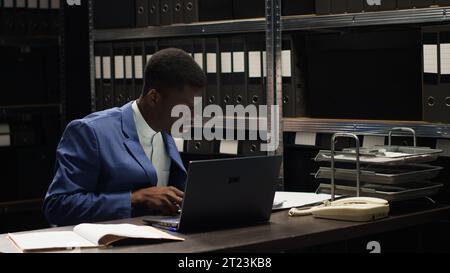 Entschlossener Privatdetektiv, der vertrauliche Dokumente aus Regalen in einer Untersuchung analysiert. Der afroamerikanische Ermittler überprüft Beweise und Aussagen auf PC und Akten. Stockfoto