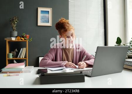 Niedliches fleißiges Schulmädchen, das auf den Laptop-Bildschirm blickt und während des Online-Unterrichts Notizen macht, während es am Schreibtisch sitzt und den Erklärungen des Lehrers zuhört Stockfoto