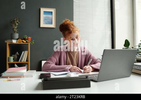 Niedliches fleißiges Schulmädchen, das auf den Laptop-Bildschirm blickt und während des Online-Unterrichts Notizen macht, während es am Schreibtisch sitzt und den Erklärungen des Lehrers zuhört Stockfoto