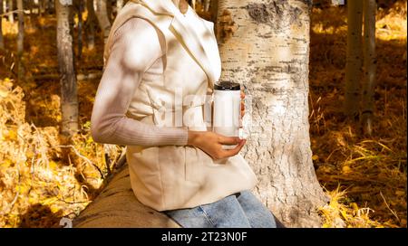 Cropped Woman Sitzt Auf Gefallenem Baum Und Trinkt Heißen Tee Von Thermos Outside In Forest, Nature, Trägt Warme Kleidung. Herbstgetränk, Herbstlaub Saison Stockfoto