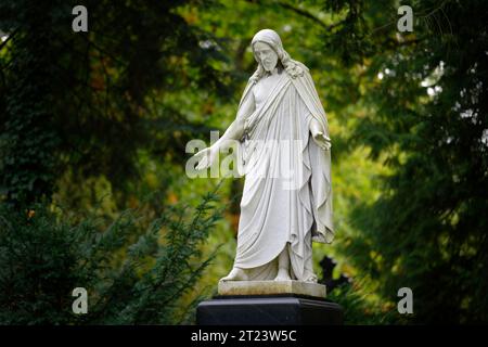 jesusfigur aus weißem Stein mit ausgebreiteten Armen, die auf einem Grabstein steht und traurig zum Boden blickt Stockfoto