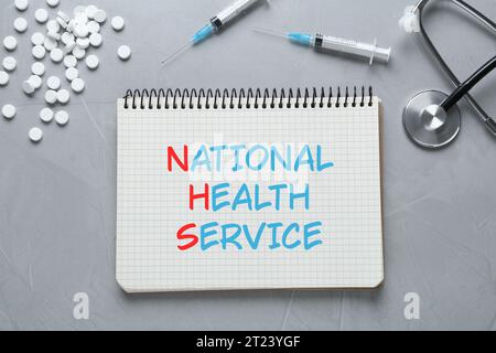 Nationaler Gesundheitsdienst (NHS). Notizbuch mit Text, Stethoskop, Pillen und Spritzen auf grauem Hintergrund, flache Auflage Stockfoto