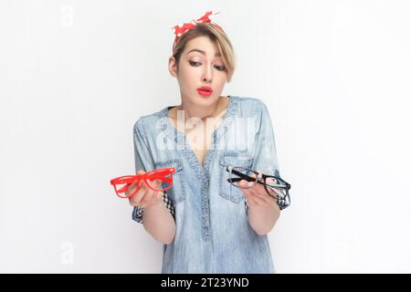 Porträt einer verwirrten blonden Frau, die blaues Jeanshemd und rotes Stirnband trägt, die rote und schwarze Brille hält, kann keine bessere Brille wählen. Innenstudio isoliert auf grauem Hintergrund. Stockfoto