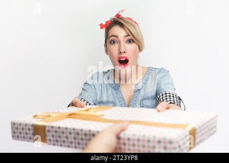 Porträt der erstaunten schockierten blonden Frau, die ein blaues Jeanshemd und ein rotes Stirnband trägt, erhält eine Geschenkbox, überrascht. Innenstudio isoliert auf grauem Hintergrund. Stockfoto