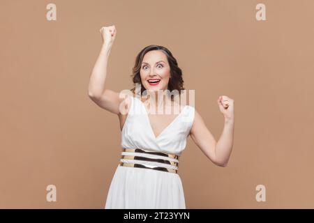 Porträt einer äußerst glücklichen Frau mittleren Alters mit gewelltem Haar, die Fäuste zusammendrückt und fröhlich schreien, ihren Triumph feiert, in weißem Kleid. Innenstudio, isoliert auf hellbraunem Hintergrund. Stockfoto
