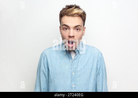 Porträt eines überraschten Mannes mit Denim-Hemd, der mit großen Augen und offenem Mund in die Kamera schaut, sieht etwas schockiertes und erstaunt. Innenstudio isoliert auf grauem Hintergrund. Stockfoto