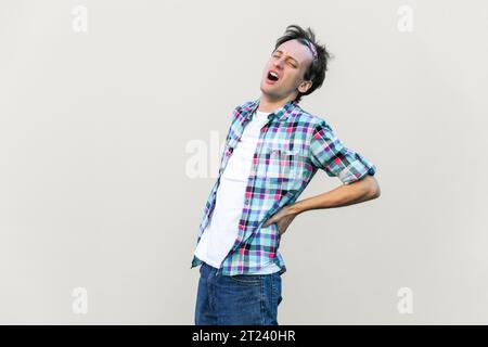 Porträt des Menschen leidet an Radikulitis, hält die Hand auf seinem schmerzhaften Rücken, krank und ungesund, trägt blaues kariertes Hemd und Stirnband. Innenstudio isoliert auf grauem Hintergrund. Stockfoto