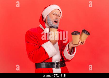 Müder, verschlafener älterer Mann mit grauem Bart, der weihnachtsmann-Kostüm trägt und viele Papierbecher mit Kaffee hält, braucht viel Energie. Innenstudio, isoliert auf rotem Hintergrund. Stockfoto