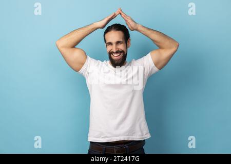 Sicherheit und Hausversicherung. Porträt eines Mannes mit Bart, der ein weißes T-Shirt trägt, der eine Geste über dem Kopf macht und mit liebenswürdigem Ausdruck zur Kamera lächelt. Innenstudio, isoliert auf blauem Hintergrund Stockfoto