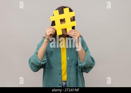 Porträt einer neugierigen Frau, die durch gelbes Hashtag-Zeichen schaut, nach richtigen Posts in sozialen Medien sucht, spioniert und eine lässige Jacke trägt. Innenstudio isoliert auf grauem Hintergrund. Stockfoto