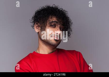 Porträt eines zuversichtlichen, stolzen, gutaussehenden bärtigen Mannes mit lockigem Haar, der mit einem Lächeln in die Kamera blickt und einen roten Pullover trägt. Innenstudio isoliert auf grauem Hintergrund. Stockfoto