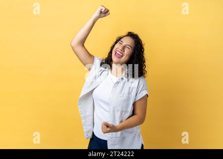 Porträt einer aufgeregten glücklichen Frau mit dunklem, welligem Haar, die gewinnende Geste mit erhobenen Fäusten und Schreien ausdrückt und den Sieg feiert. Innenstudio, isoliert auf gelbem Hintergrund. Stockfoto