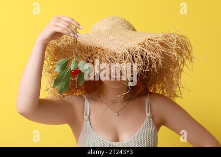 Glückliche schöne junge Frau im Weidenhut mit Zweig reifer Kirschen auf gelbem Hintergrund Stockfoto