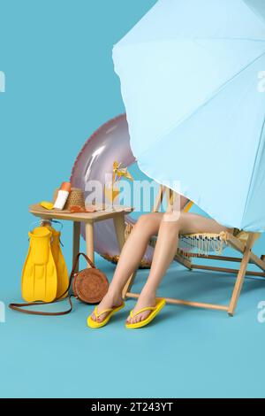 Junge Frau, die im Liegestuhl mit einem Glas Cocktail und Strandaccessoires auf blauem Hintergrund sitzt Stockfoto