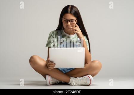 Interessiertes Teenager-Mädchen in Brille, die Lektion auf Laptop auf grauem Hintergrund ansieht. Stockfoto