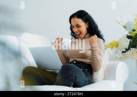 Erfolgreiche afroamerikanische Geschäftsfrau fühlt sich glücklich über die entscheidende Arbeit am Laptop, während sie zu Hause auf der Sofaecke sitzt Stockfoto