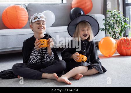 Niedliche Geschwister in Karnevalskostümen sitzen auf dem Boden und haben Spaß auf der Halloween-Party Stockfoto