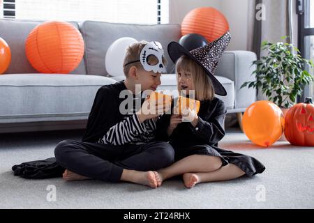 Niedliche Geschwister in Karnevalskostümen sitzen auf dem Boden und haben Spaß auf der Halloween-Party Stockfoto