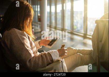 Unerkennbare Frau, die Smartphone benutzt und beim Essen im Wartezimmer des Flughafens mit Gepäck sitzt Stockfoto