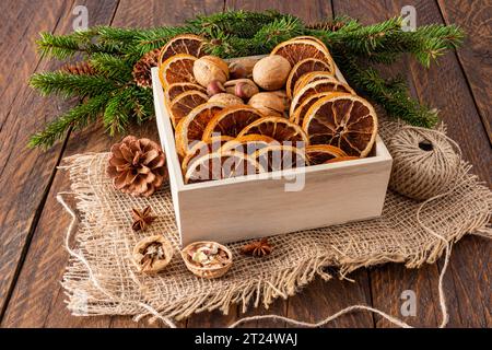 Quadratische Holzkiste mit getrockneten Orangenscheiben und Walnüssen auf Sackleinen und rustikaler Tisch mit grünen Fichtenzweigen. Umweltfreundlicher Weihnachtsstil Stockfoto