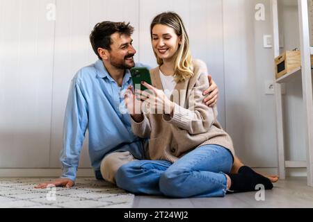 Ein junges glückliches Paar, das zu Hause Nachrichten aus sozialen Medien per Telefon mitteilt und online einkauft. Stockfoto
