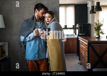 Fröhliches, junges Paar, bedeckt mit einer Decke, die sich umschließt, während man zu Hause Tassen Tee hält Stockfoto