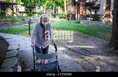 Eine ältere Frau mit einer Gesichtsmaske, die draußen läuft, schiebt den Mobilitätsläufer in einem Pensionsdorf. Stockfoto