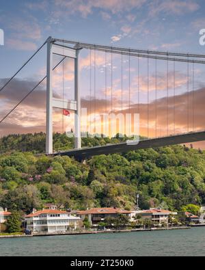 Blick vom Meer auf die asiatische Seite der Bosporus Meerenge, Istanbul, Türkei, mit traditionellen Häusern am Wasser, unter der Bosporus-Brücke und grünem Hügel mit dichten Bäumen vor Sonnenuntergang Stockfoto