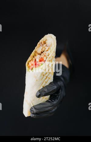 Hand hält Döner oder Shawarma durch gerissene Löcher auf schwarzem Papierhintergrund. Street-Food-Konzept Stockfoto