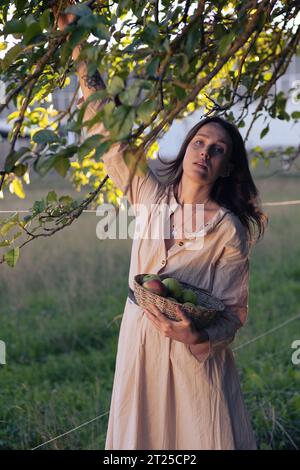 Schönes Mädchen im Apfelgarten. Eine junge Frau in einem schönen hellen Kleid sammelt grüne Äpfel in einem Korb zwischen Apfelbäumen. Ernte A Stockfoto