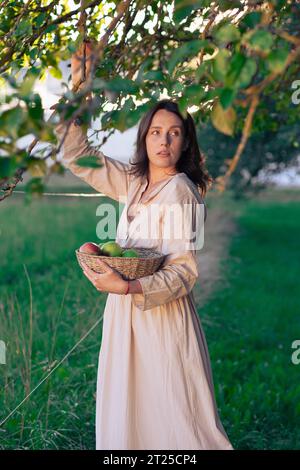 Schönes Mädchen im Apfelgarten. Eine junge Frau in einem schönen hellen Kleid sammelt grüne Äpfel in einem Korb zwischen Apfelbäumen. Ernte A Stockfoto